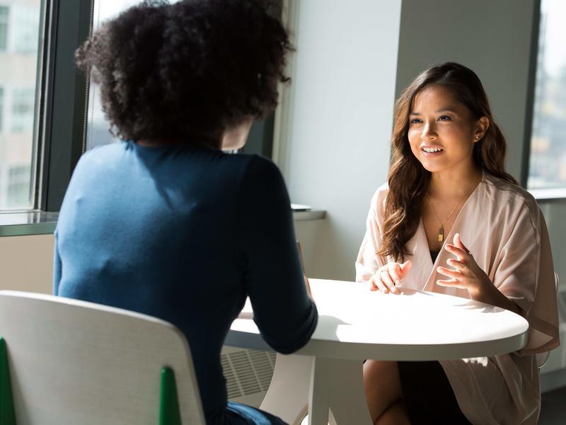 Two women wearing business attire having a discussion at a white table in front of a window.